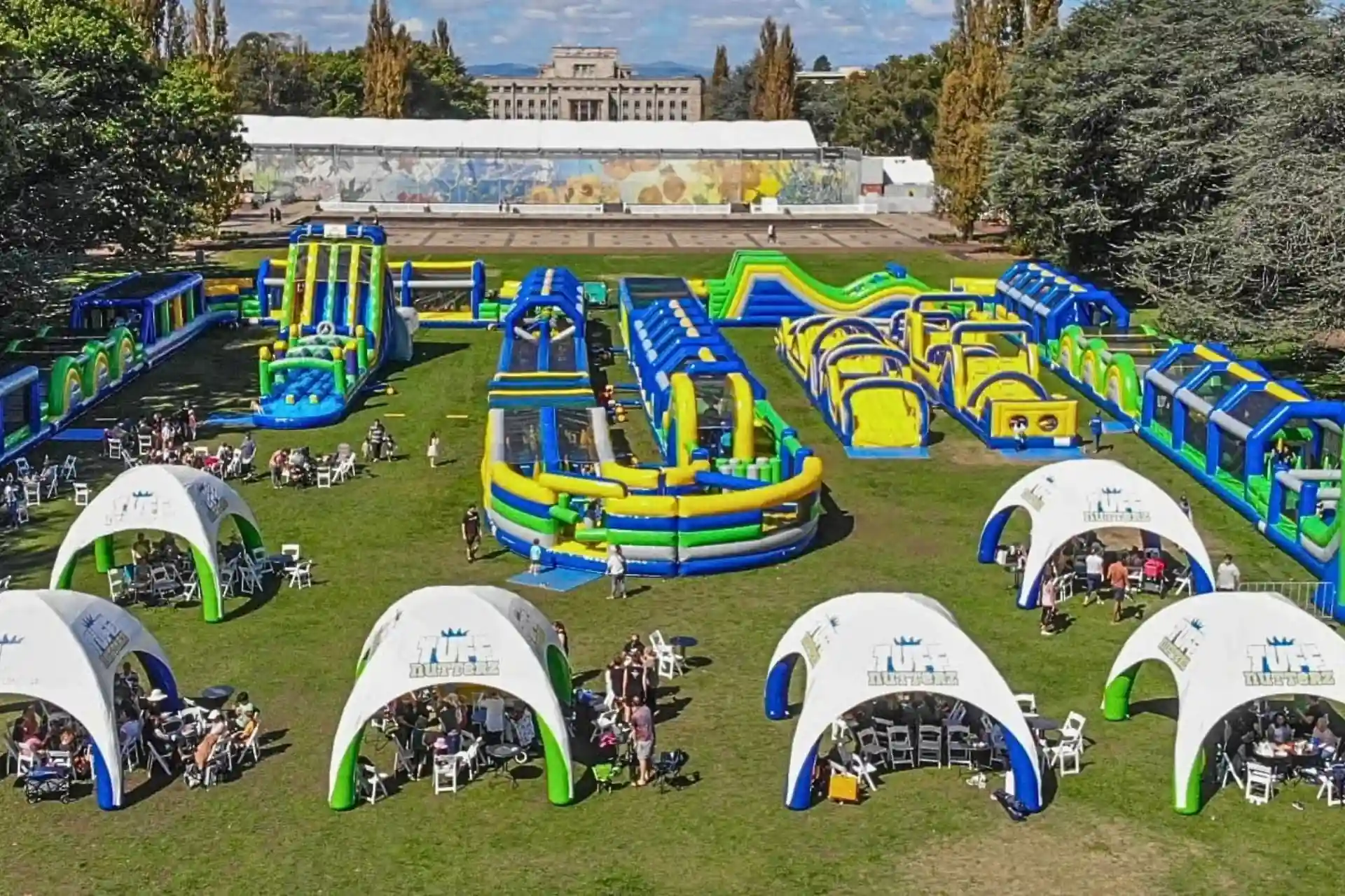 Aerial view of a massive blue, green, and yellow Tuff Nutterz inflatable obstacle course set up on a grassy lawn with white event tents in the foreground.