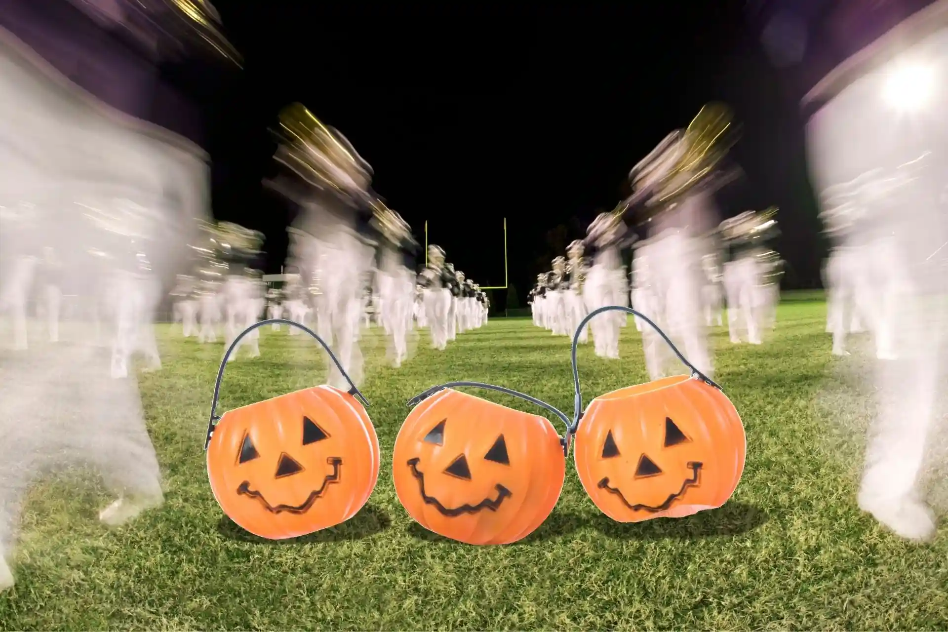 Three orange jack-o'-lantern trick-or-treat buckets in the foreground of a marching band performing on a football field at night.