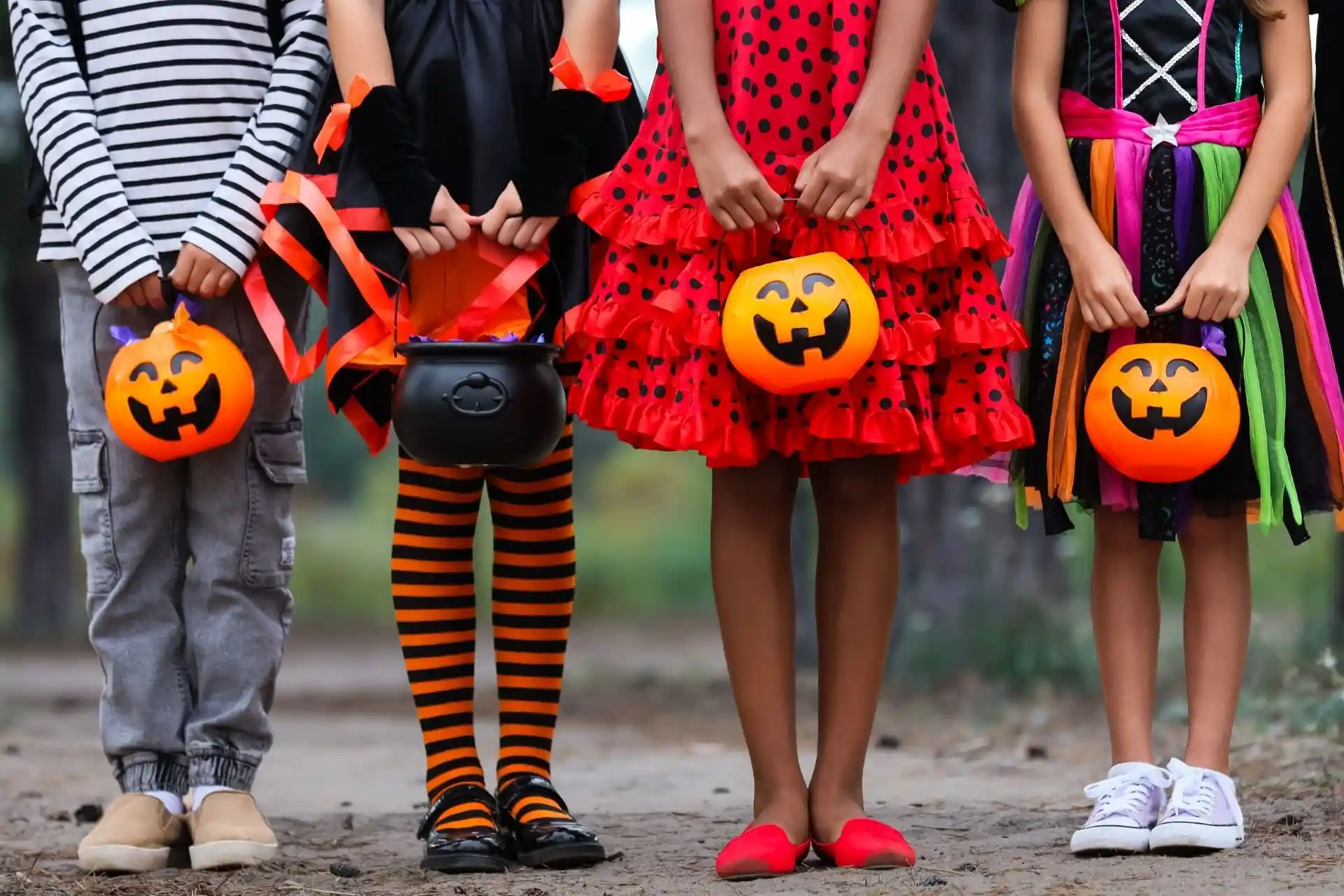 Four children in colorful Halloween costumes holding pumpkin-shaped candy buckets and a black cauldron while standing outdoors.