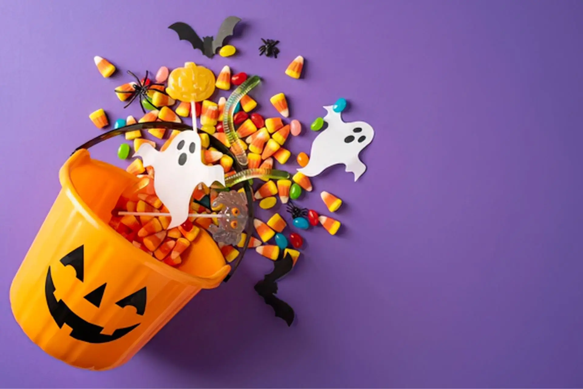 Orange jack-o'-lantern trick-or-treat bucket spilling candy corn, gummy worms and Halloween decorations onto a purple background.