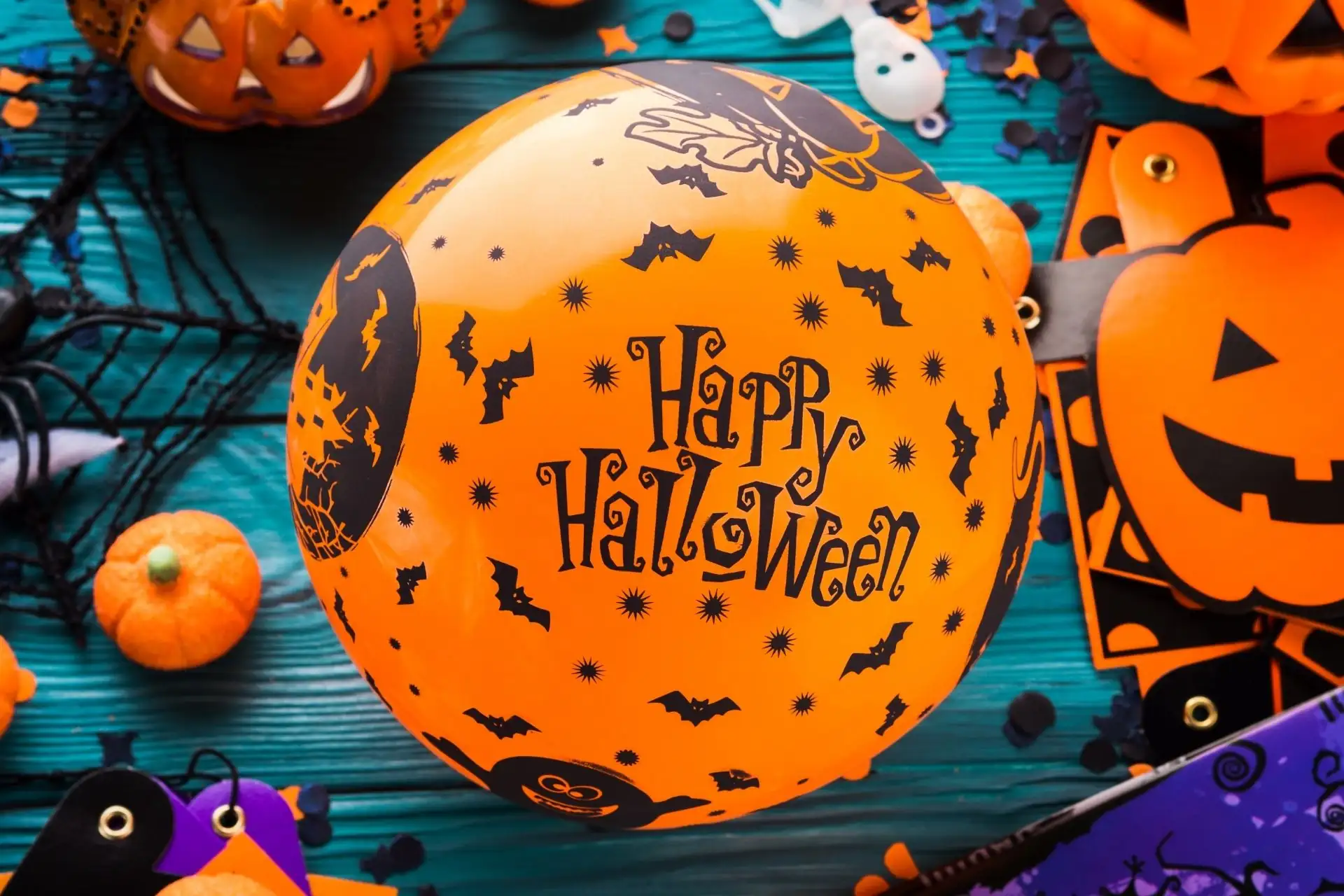 Orange Happy Halloween balloon decorated with black bats, surrounded by pumpkins, jack-o'-lantern decorations and Halloween party supplies on a turquoise wooden background.