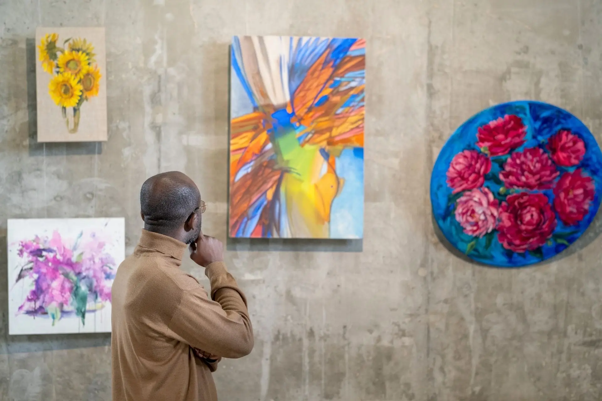 A visitor admiring colorful floral and abstract oil paintings displayed on a concrete wall inside an art gallery.