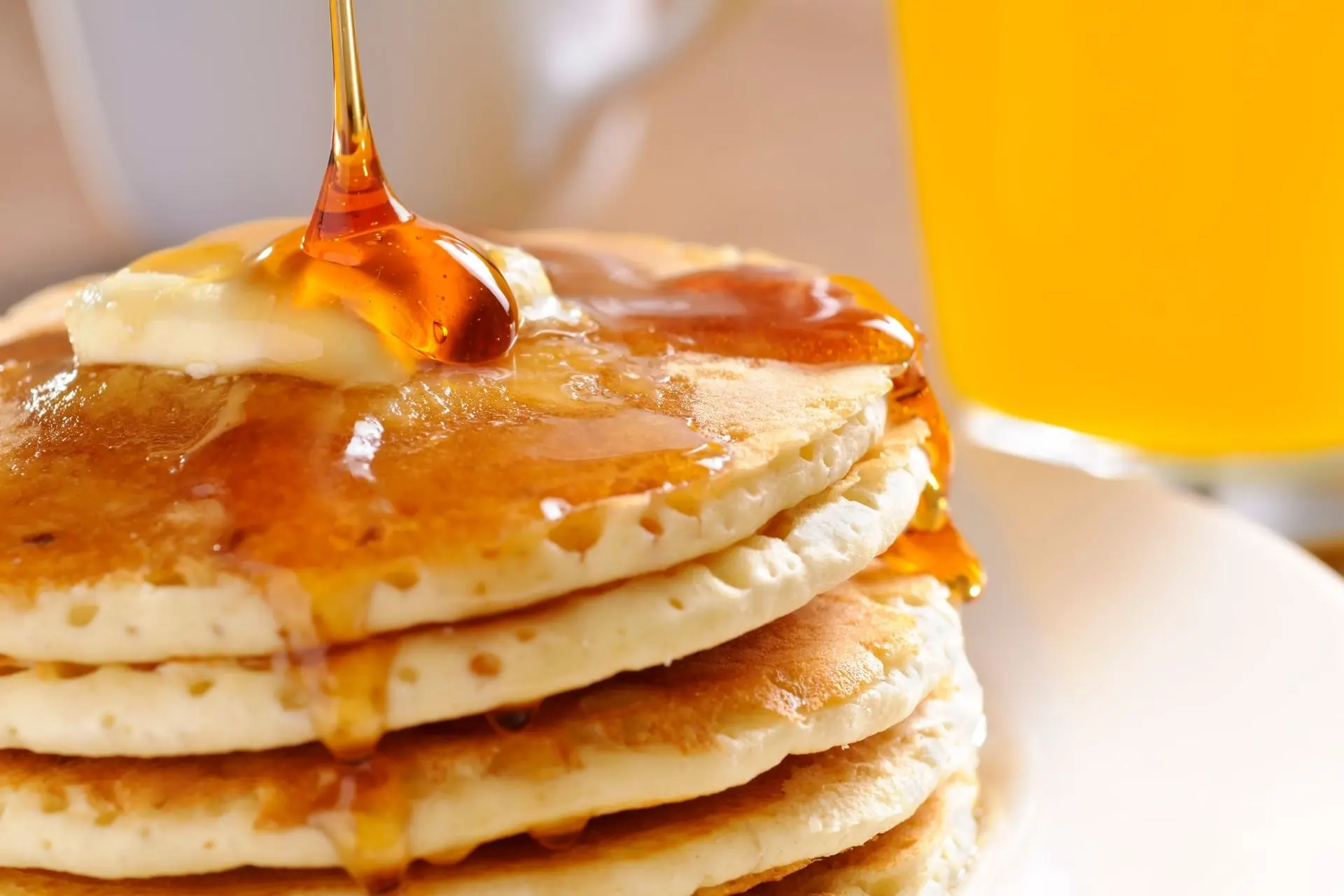A stack of pancakes on a plate with a pat of butter on top and maple syrup being poured over it.