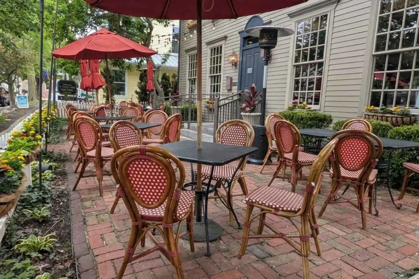An outdoor dining patio on a brick sidewalk in downtown Zionsville with red umbrellas and woven bistro chairs next to a historic building.