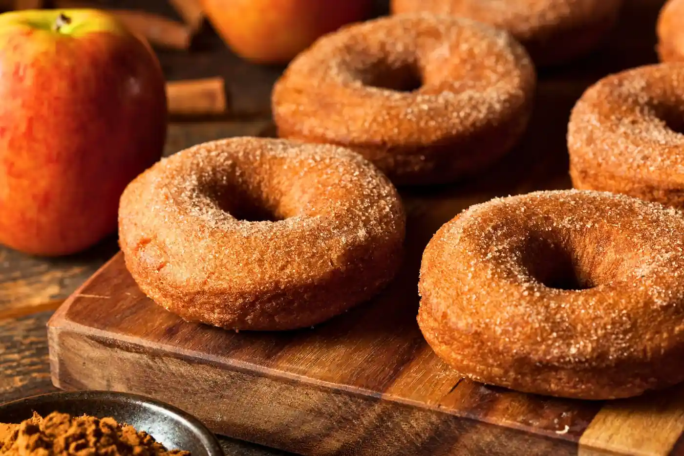 Apple cider donuts coated in cinnamon sugar sitting on a wooden board next to a red apple.