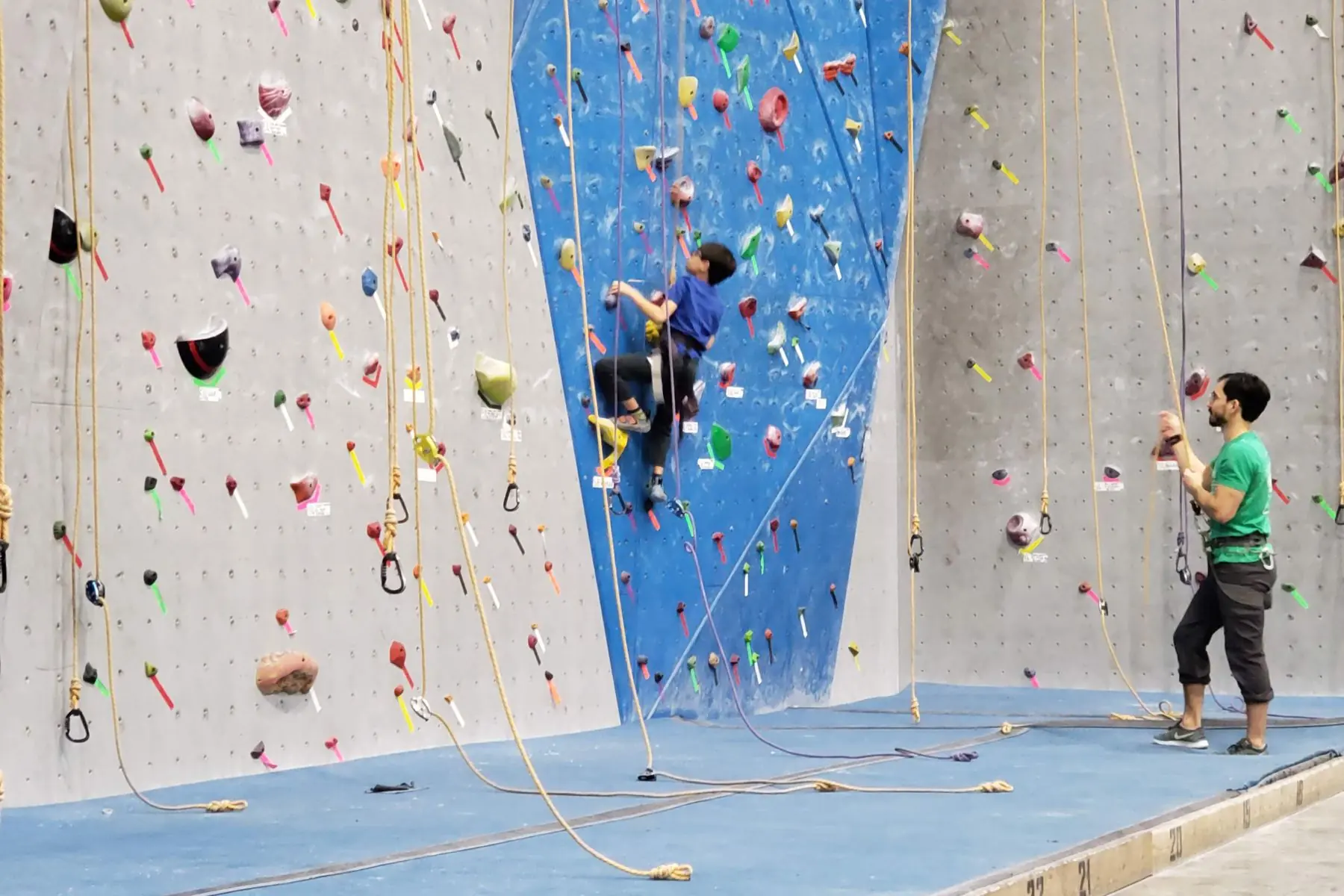 Child climbing a colorful indoor rock wall at Hoosier Heights in Carmel, Indiana, with an adult nearby.