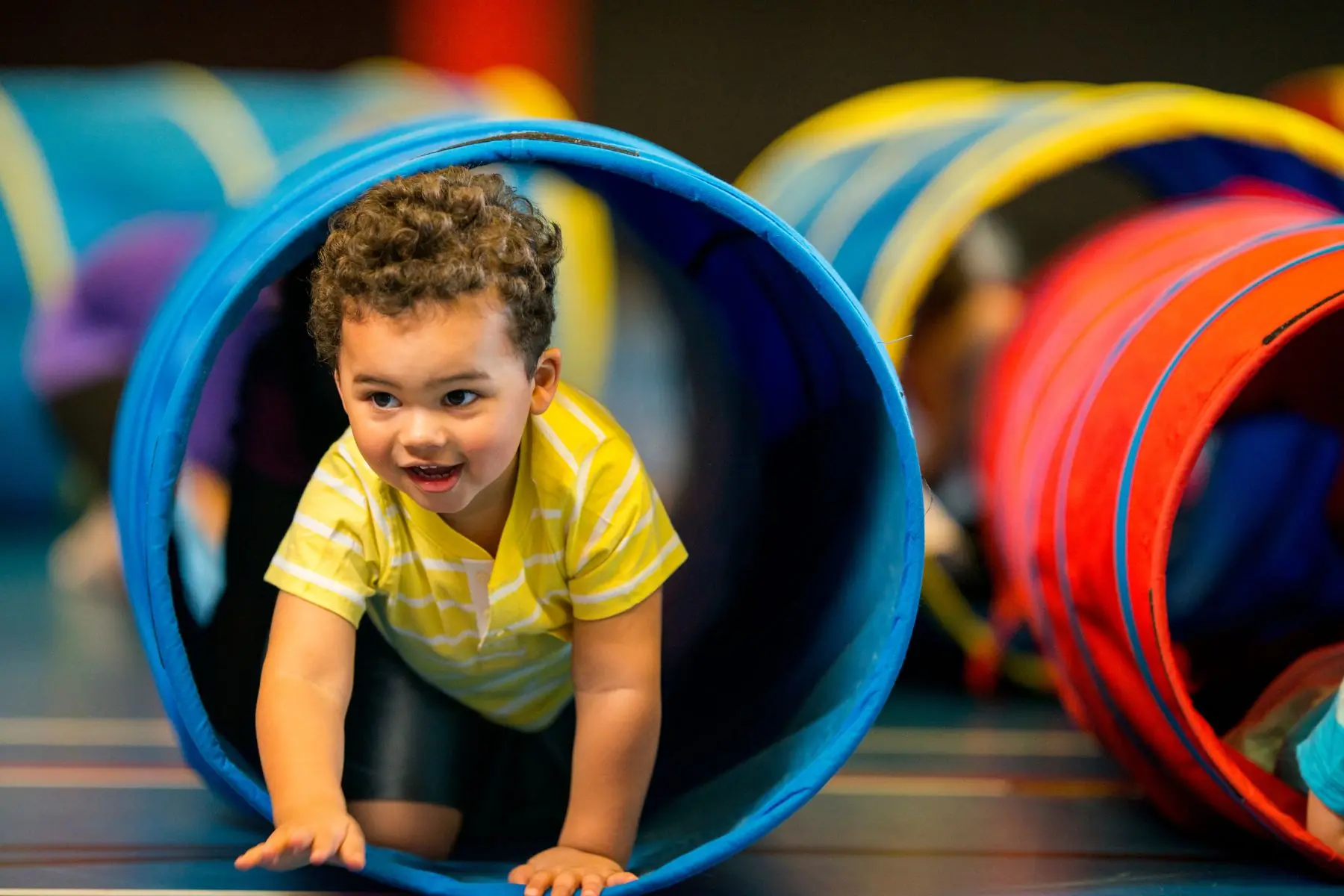 A young toddler crawling through a blue fabric play tunnel in an indoor gymnastics and obstacle play area.