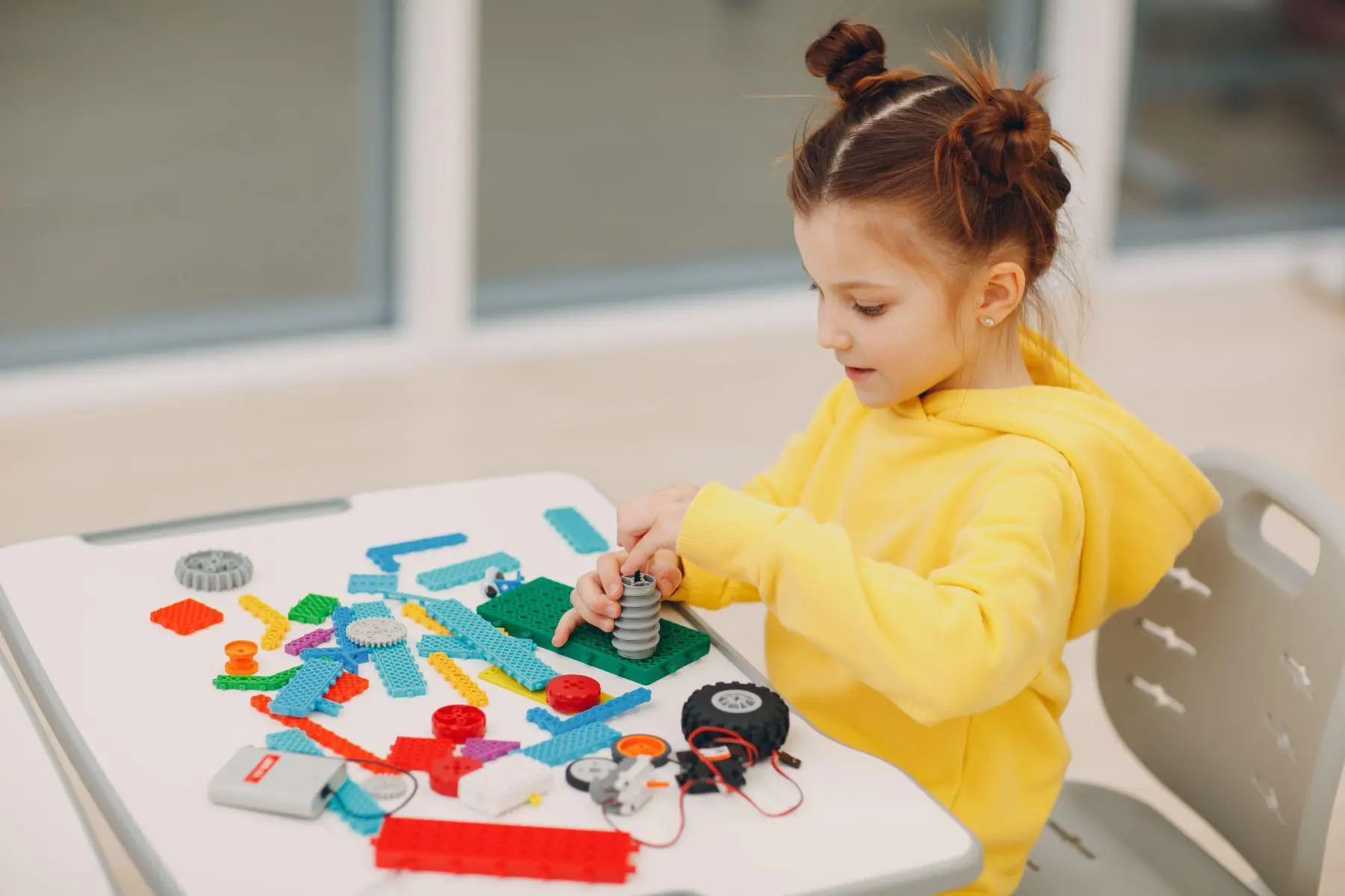 Young girl assembling a robot with colorful building pieces, gears, wheels, and electronic components during a STEM activity.