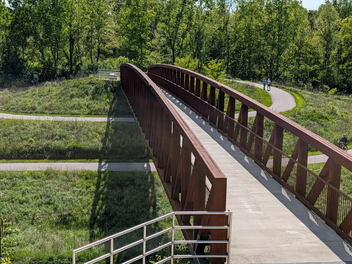 A rusty-brown steel truss pedestrian bridge with stairs leading down to paved multi-use paths that loop through the grassy fields of Overley-Worman Park in Zionsville, Indiana.