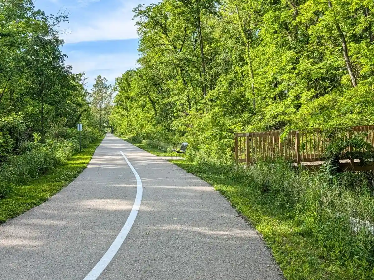 A straight paved section of the Big-4 Rail Trail in Zionsville, Indiana, featuring a white centerline, a wooden pedestrian bridge on the right, and a park bench alongside the wooded path.