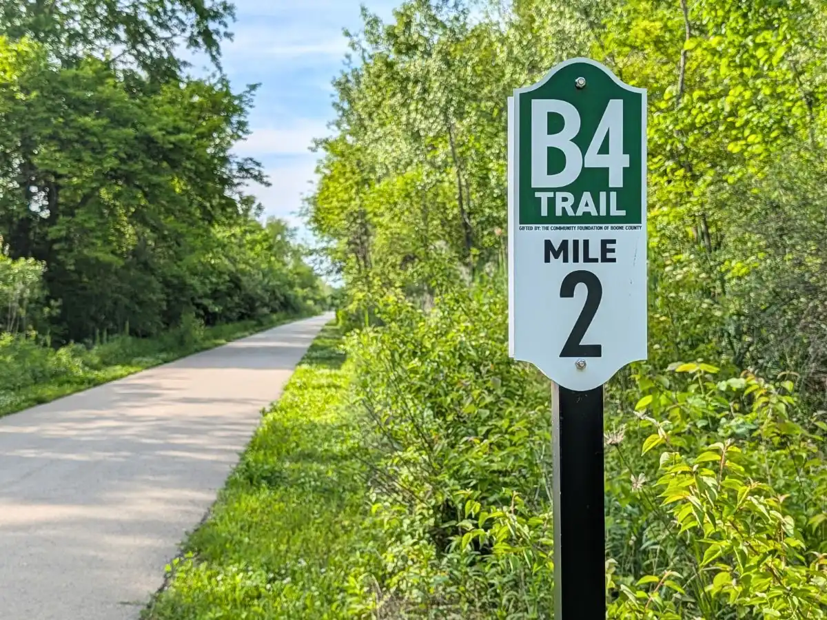 A green and white metal sign reading "B4 TRAIL MILE 2" mounted on a black post along the edge of the paved Big-4 Rail Trail, surrounded by dense green bushes and trees.