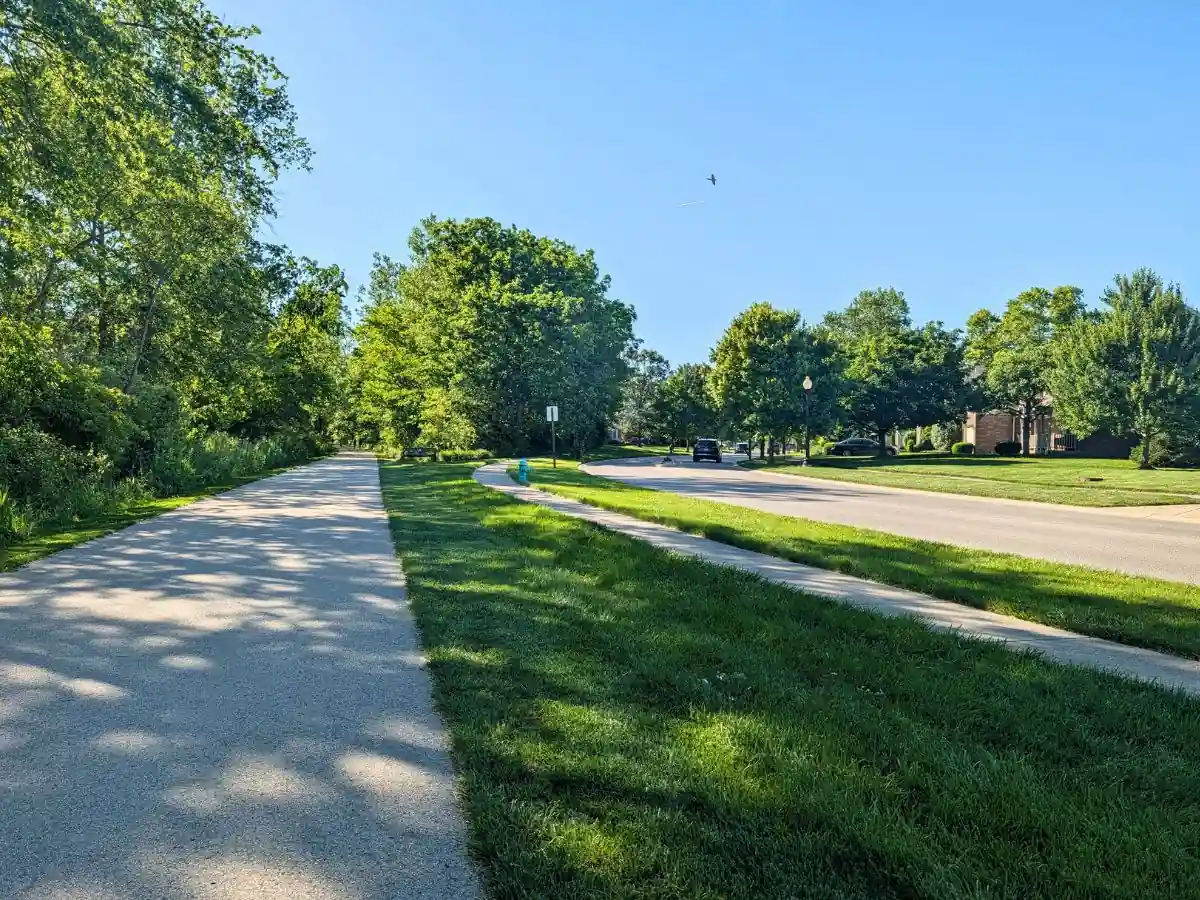 A wide, paved section of the Big-4 Rail Trail running parallel to a grassy area with a blue fire hydrant, a sidewalk, and a quiet residential neighborhood street lined with mature trees.