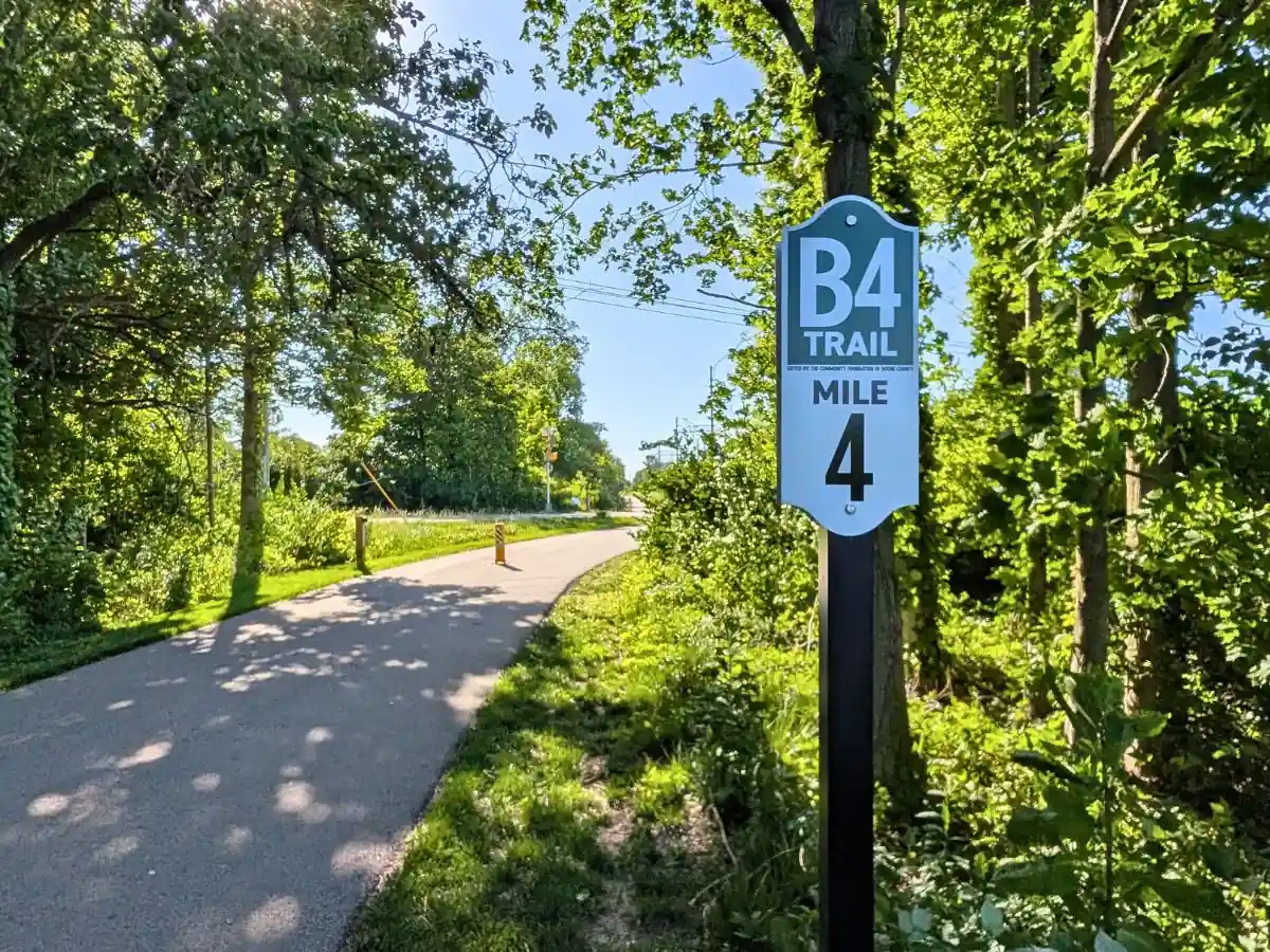 A green and white trail marker sign reading "B4 TRAIL MILE 4" on a black post next to a paved asphalt path that curves gently through a wooded area under a clear sky.