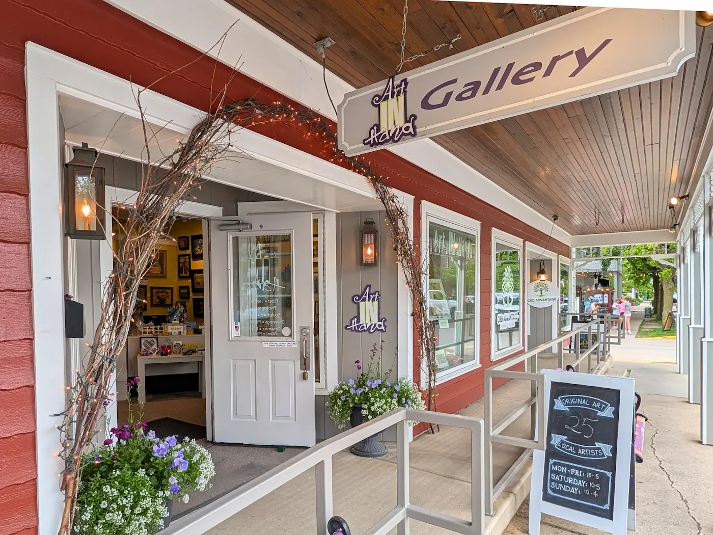 The storefront entrance and accessibility ramp of Art IN Hand Gallery on Main Street in downtown Zionsville, featuring an overhead wooden sign and a sidewalk chalkboard display.