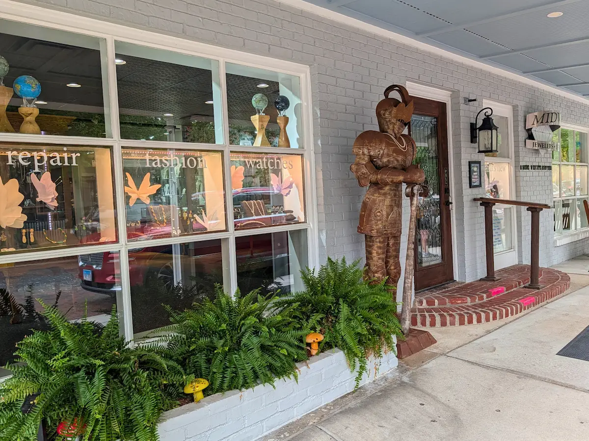 The storefront window and main entryway of Midwest Jewelers & Estate Buyers in downtown Zionsville, featuring a life-size metal suit of armor statue and a brick planter filled with ferns.