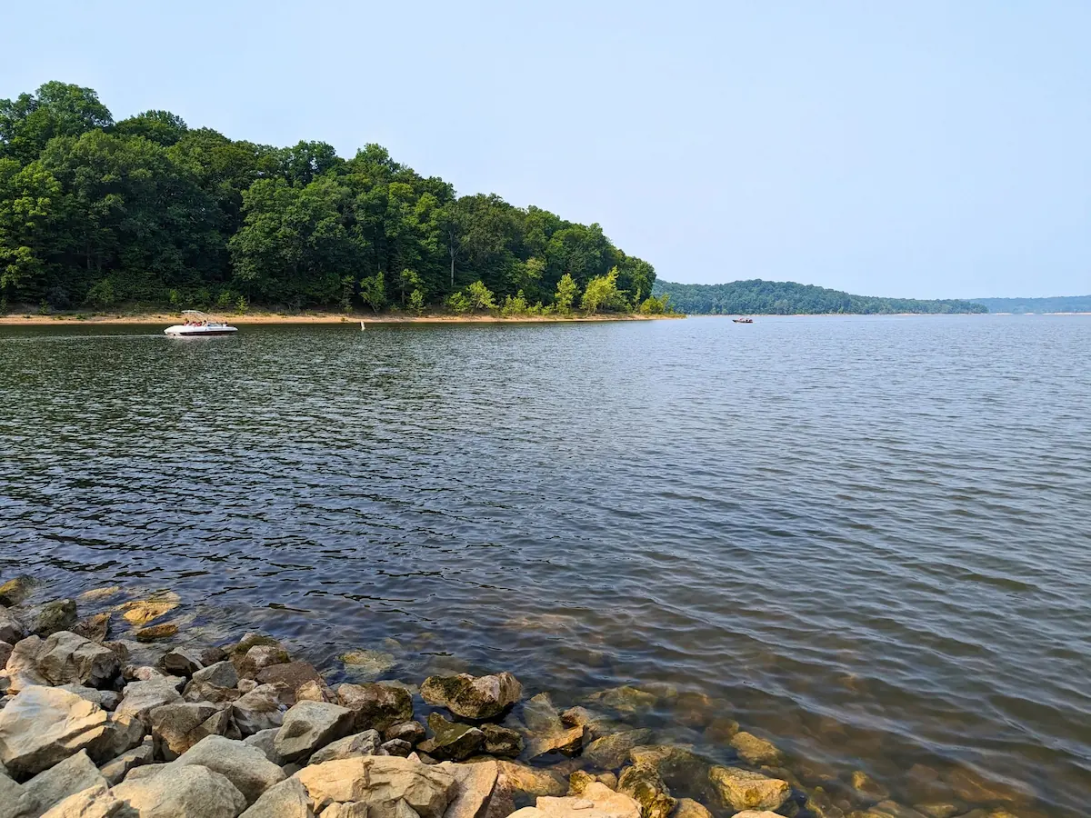 A scenic view of Lake Monroe in Indiana on a clear summer day, featuring a rocky shoreline in the foreground, a pontoon boat on the water, and a dense green forest lining the distant shore.