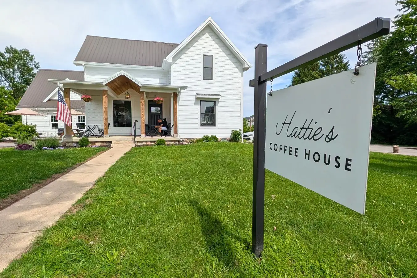 Hattie's Coffee House in Whitestown, shown in a renovated white farmhouse with a front porch, hanging flower baskets, lawn, sidewalk, and roadside sign.