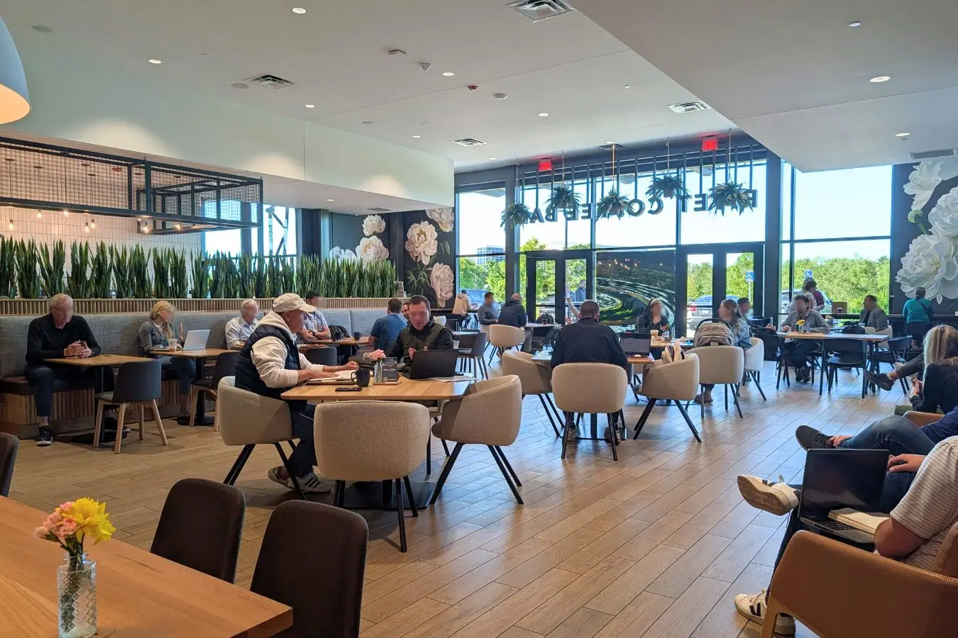 RIVET Coffee Bar in Zionsville, showing a large modern seating area with café tables, lounge chairs, tall windows, hanging plants, floral wall art, and a motorsports display.