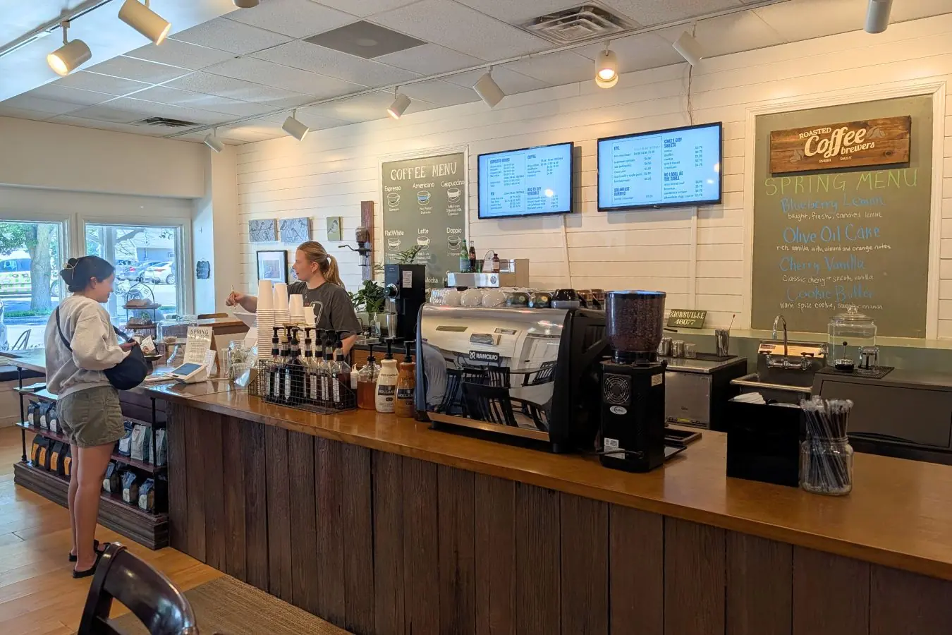 Roasted in the Village coffee counter in downtown Zionsville, with a wood service bar, espresso machine, bottled syrups, menu boards, and a spring drink menu.
