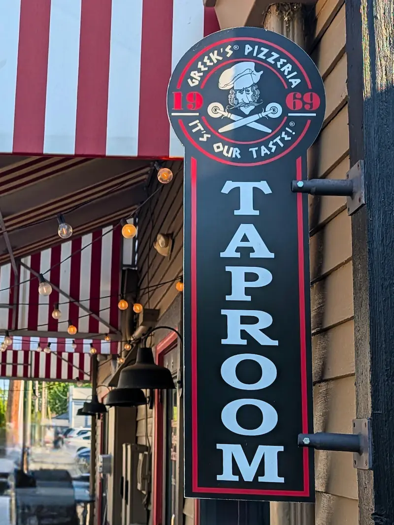 The exterior hanging sign for Greek's Pizzeria Taproom, positioned beneath a red and white striped awning with warm cafe string lights.