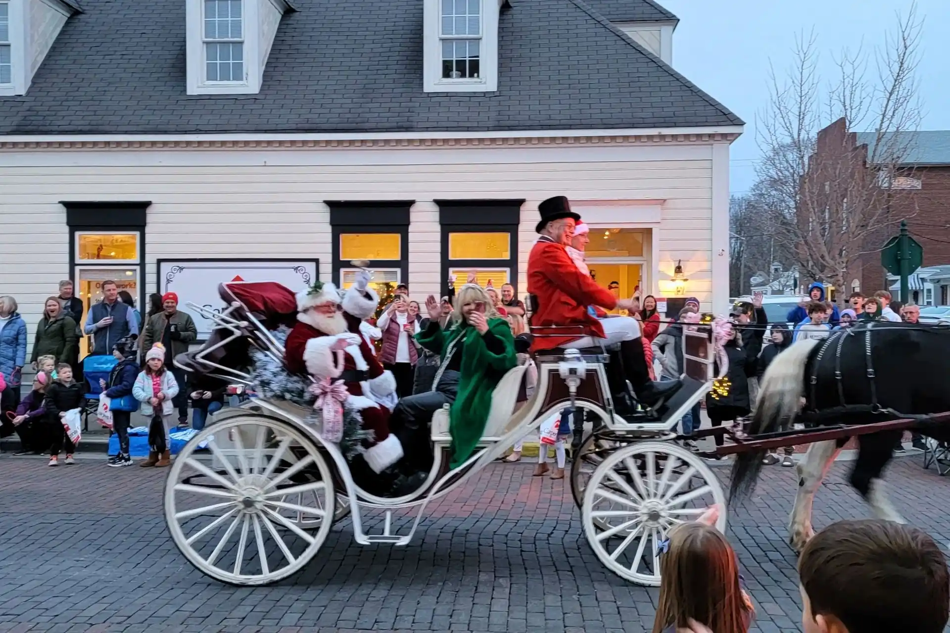 Santa Claus and Mrs. Claus waving from a horse-drawn carriage during the holiday parade along brick Main Street in Zionsville, Indiana.