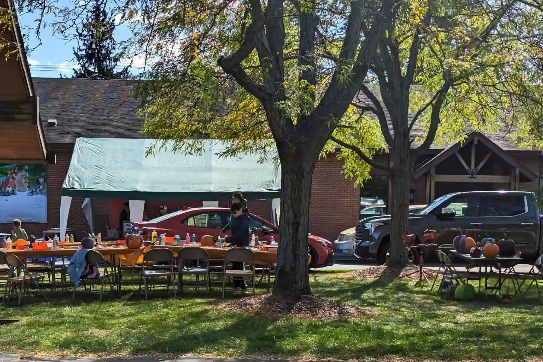 Outdoor pumpkin decorating station set under shade trees with long tables holding paint bottles, brushes, chairs, and a smaller display table on the right with painted pumpkins drying.