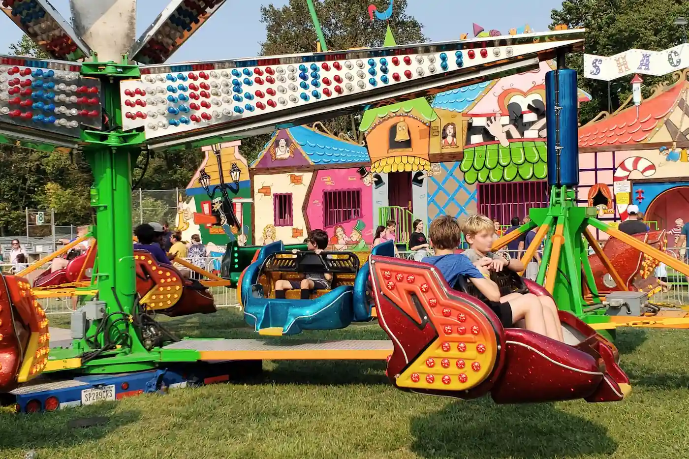 Children riding colorful spinning carnival swings against a backdrop of festive funhouse facades.