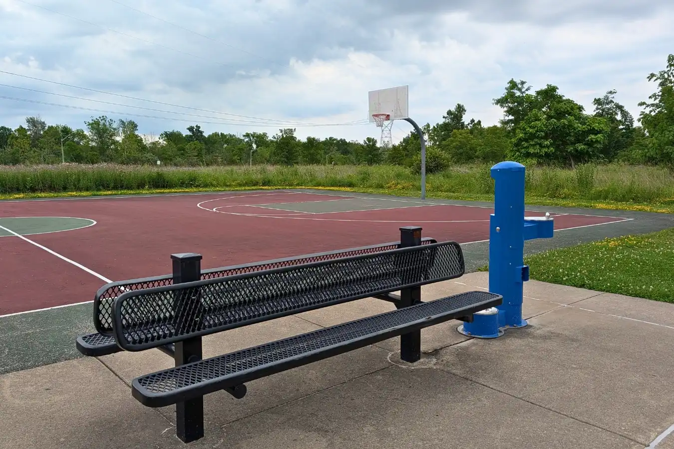 An outdoor basketball court at Mulberry Fields Park in Zionsville, featuring a painted red and green playing surface, a single hoop, a black metal mesh bench, and a blue drinking fountain.