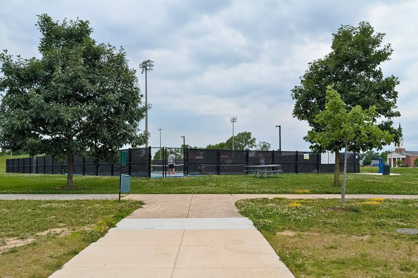 A concrete pathway leading toward the fenced outdoor pickleball courts at Mulberry Fields Park in Zionsville, featuring black windscreens on the fence, surrounding grass, and trees.