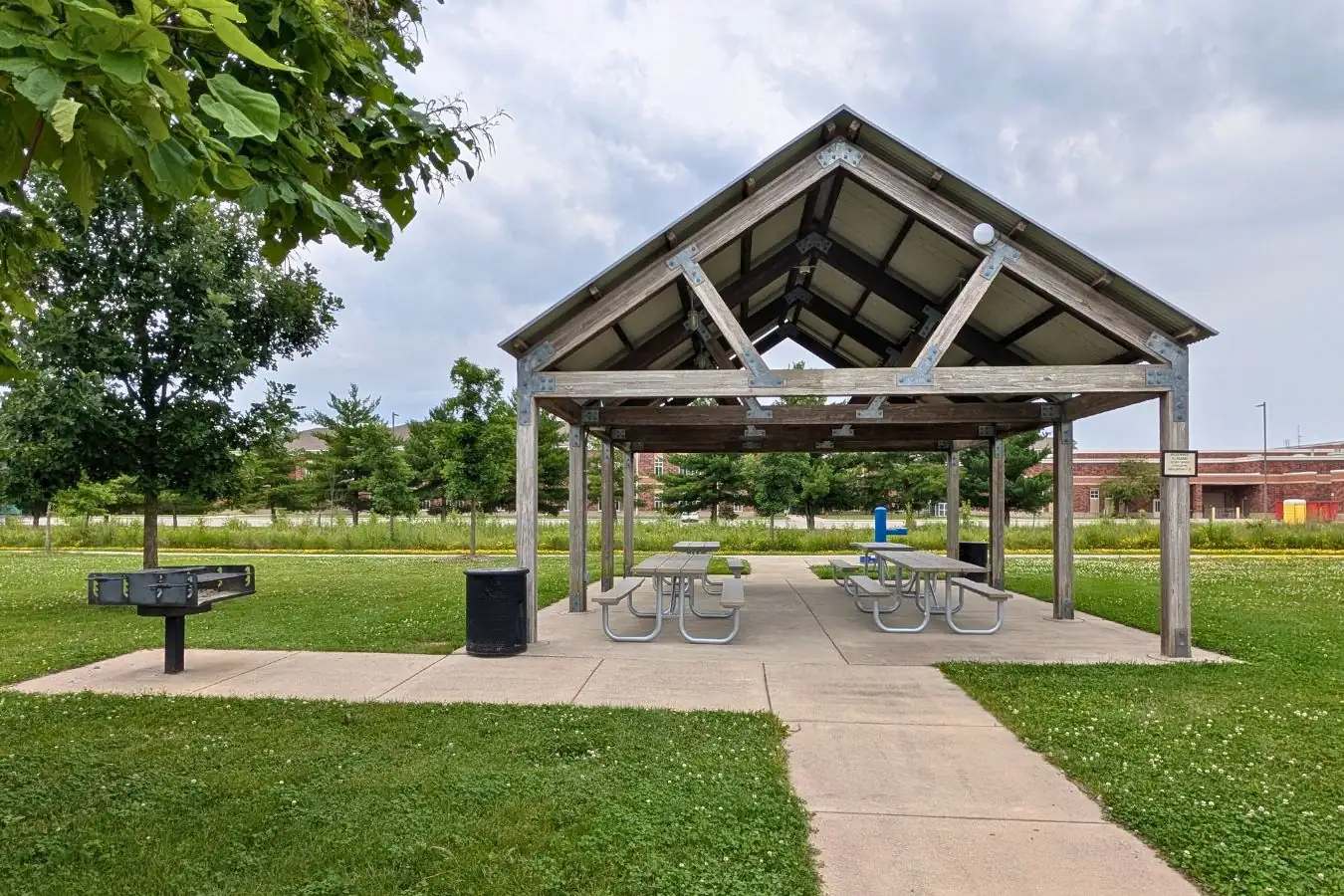 An open-air wooden timber frame picnic pavilion with a metal roof and picnic tables on a concrete pad at Mulberry Fields Park, featuring a charcoal grill on the left.